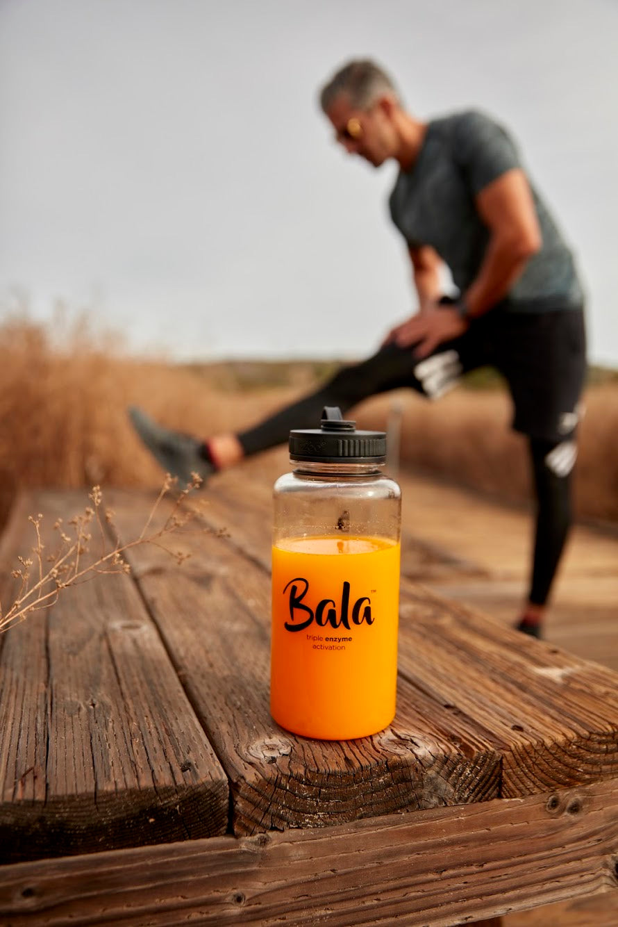 A 34oz bottle of The Bala Bottle by Bala Enzyme, a BPA-free Total Body Wellness Drink, sits on a wooden surface while a person in athletic attire stretches in the background.