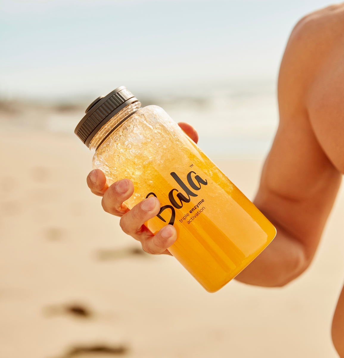 A person holding a 34oz bottle of "The Bala Bottle" from Bala Enzyme with an orange beverage on a sunny beach, the clear, BPA-free container catching the sunlight.